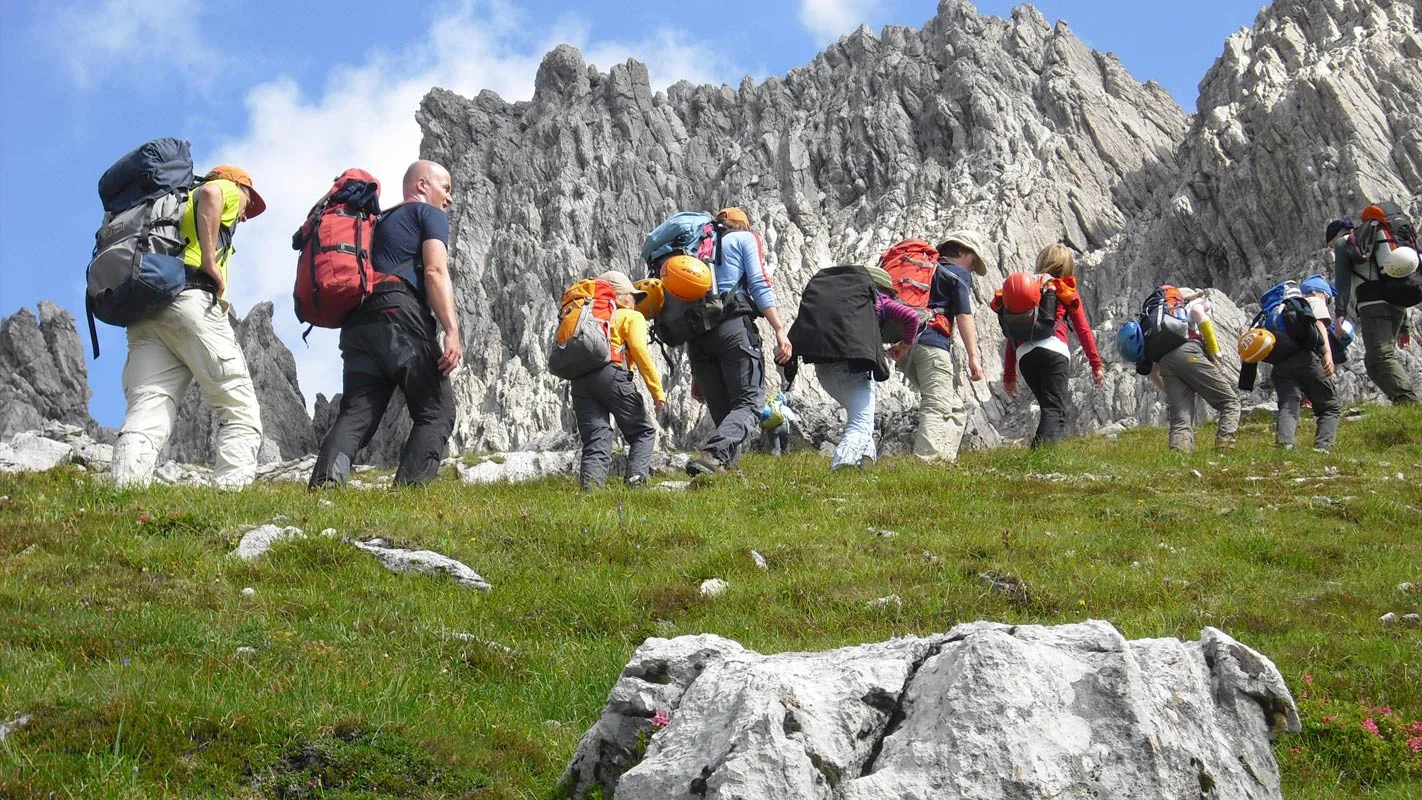 Es wird gewandert bei sonnigen Wetter auf einer Art Wiese, Im Hintergrund steile Felswände | © DAV Münster