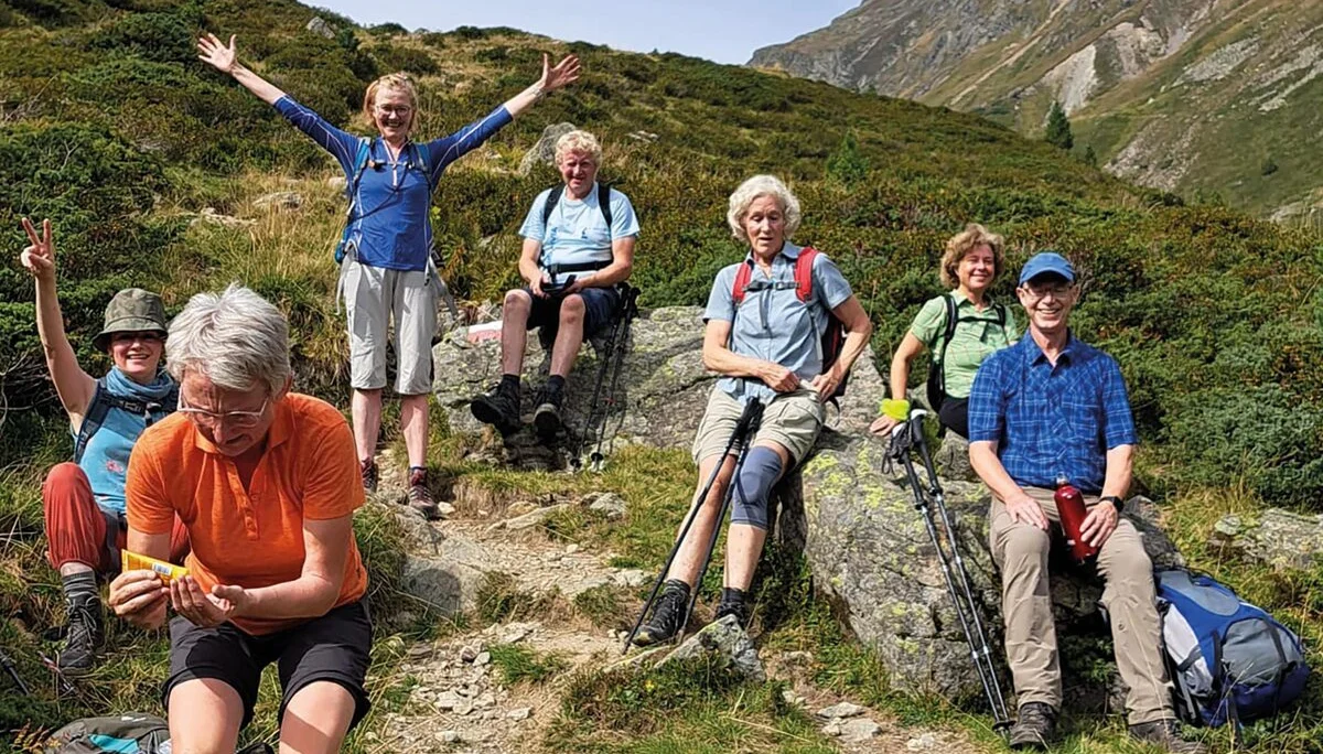 Es sind sieben Wanderer in den Bergen zu sehen die eine Pause machen.  Sechs sitzen z.B. auf Steinen, eine steht aufrecht und hat die Hände nach oben gestreckt. Im Hintergrund sind Berge zu erahnen.  | © DAV Münster