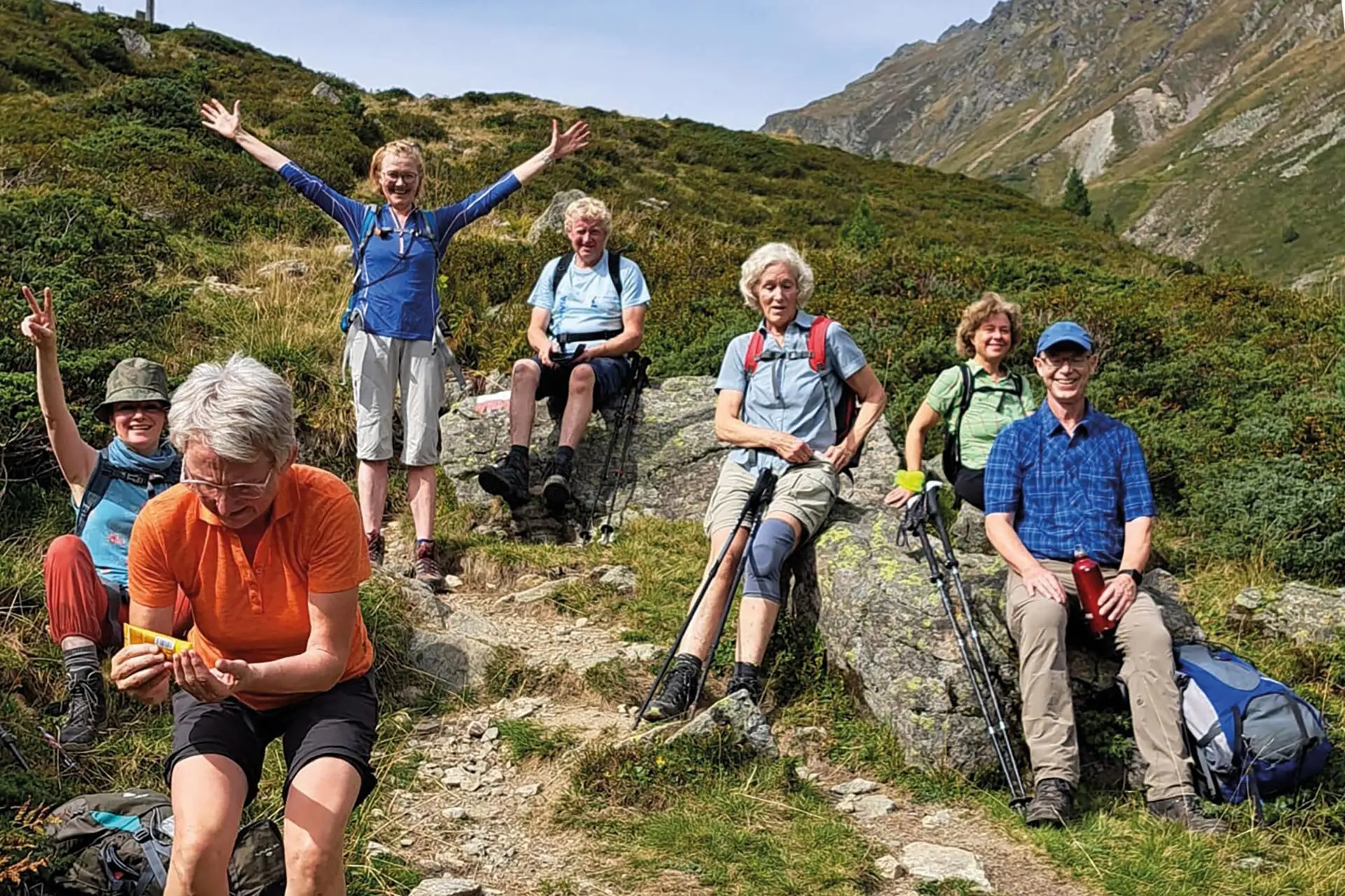 Es sind sieben Wanderer in den Bergen zu sehen die eine Pause machen.  Sechs sitzen z.B. auf Steinen, eine steht aufrecht und hat die Hände nach oben gestreckt. Im Hintergrund sind Berge zu erahnen.  | © DAV Münster