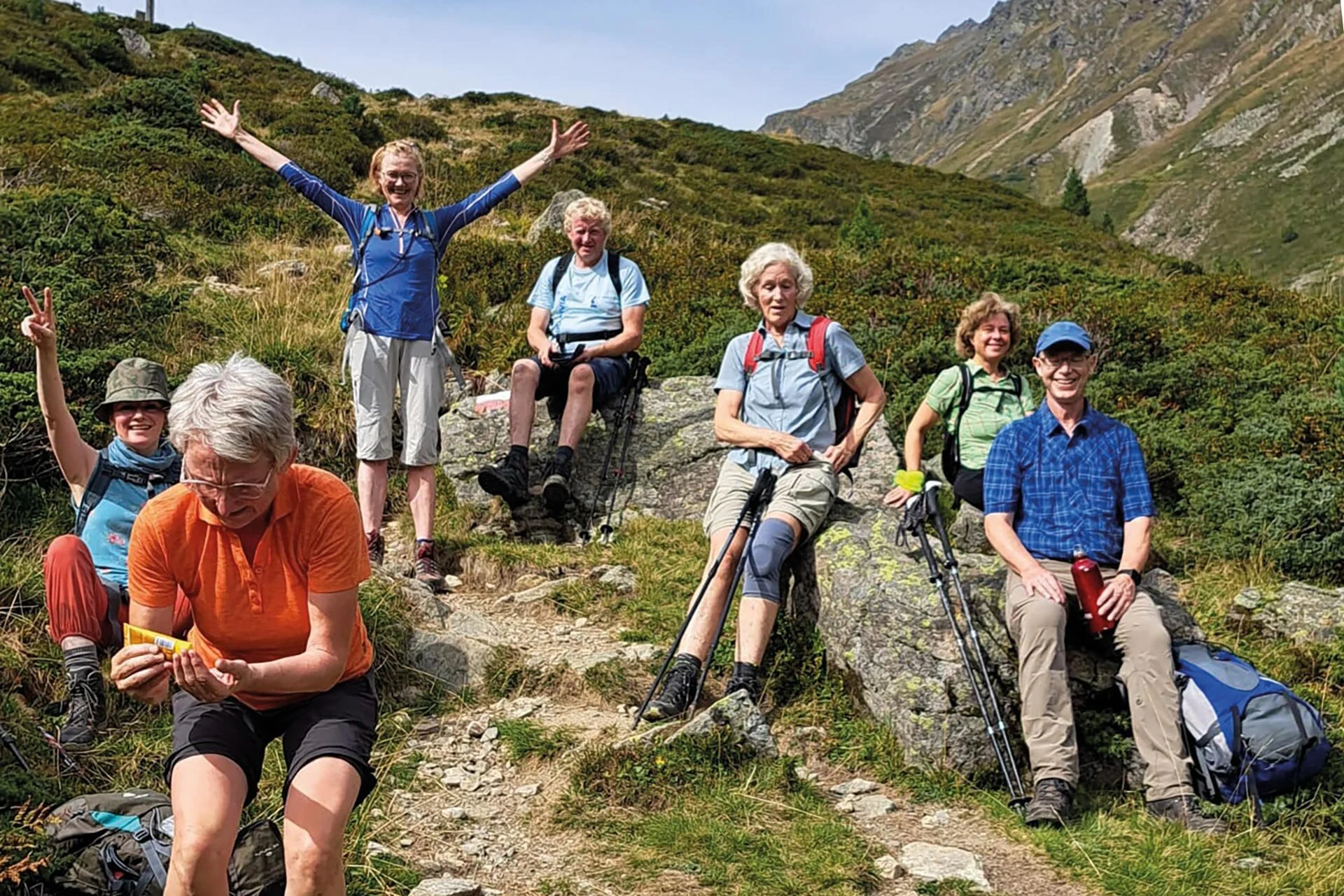 Es sind sieben Wanderer in den Bergen zu sehen die eine Pause machen.  Sechs sitzen z.B. auf Steinen, eine steht aufrecht und hat die Hände nach oben gestreckt. Im Hintergrund sind Berge zu erahnen.  | © DAV Münster