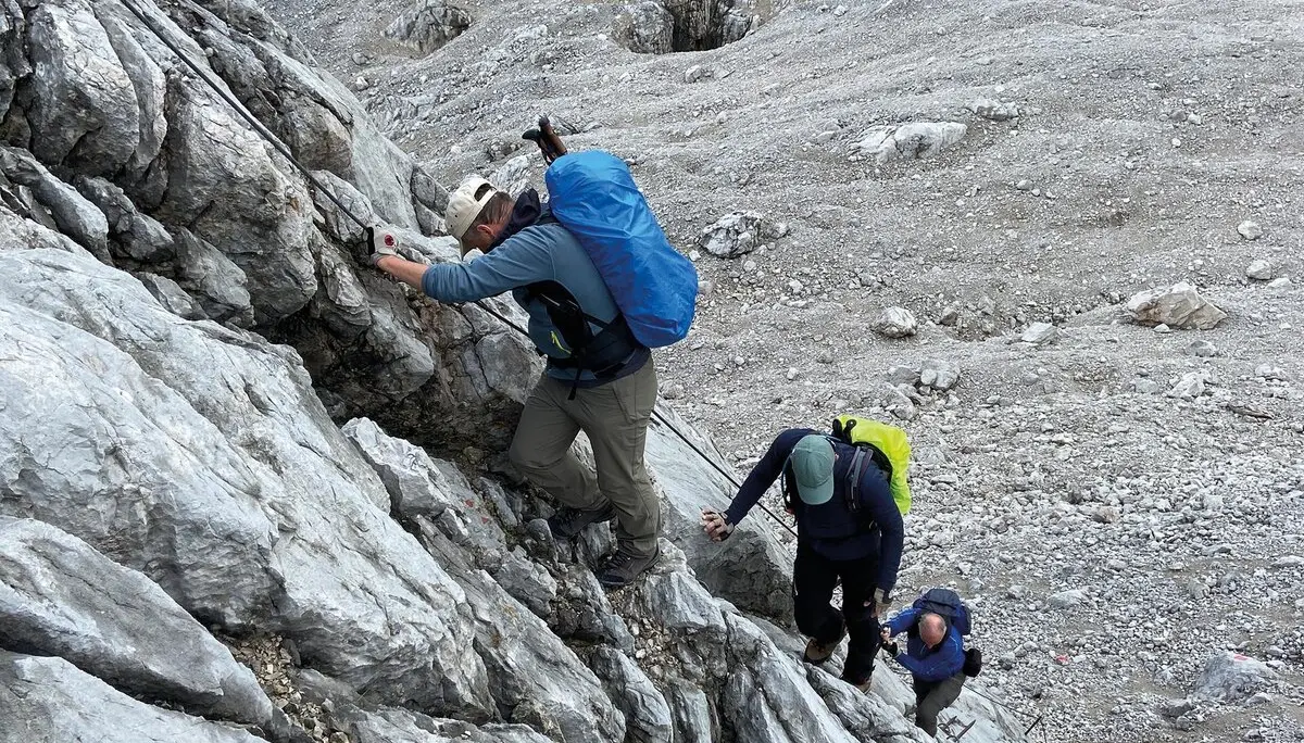 Es sind drei Wanderer mit Rucksäcken zu sehen, die an einem Sicherungsseil nach oben klettern. Alle schauen nach unten oder zur Seite. | © DAV Münster
