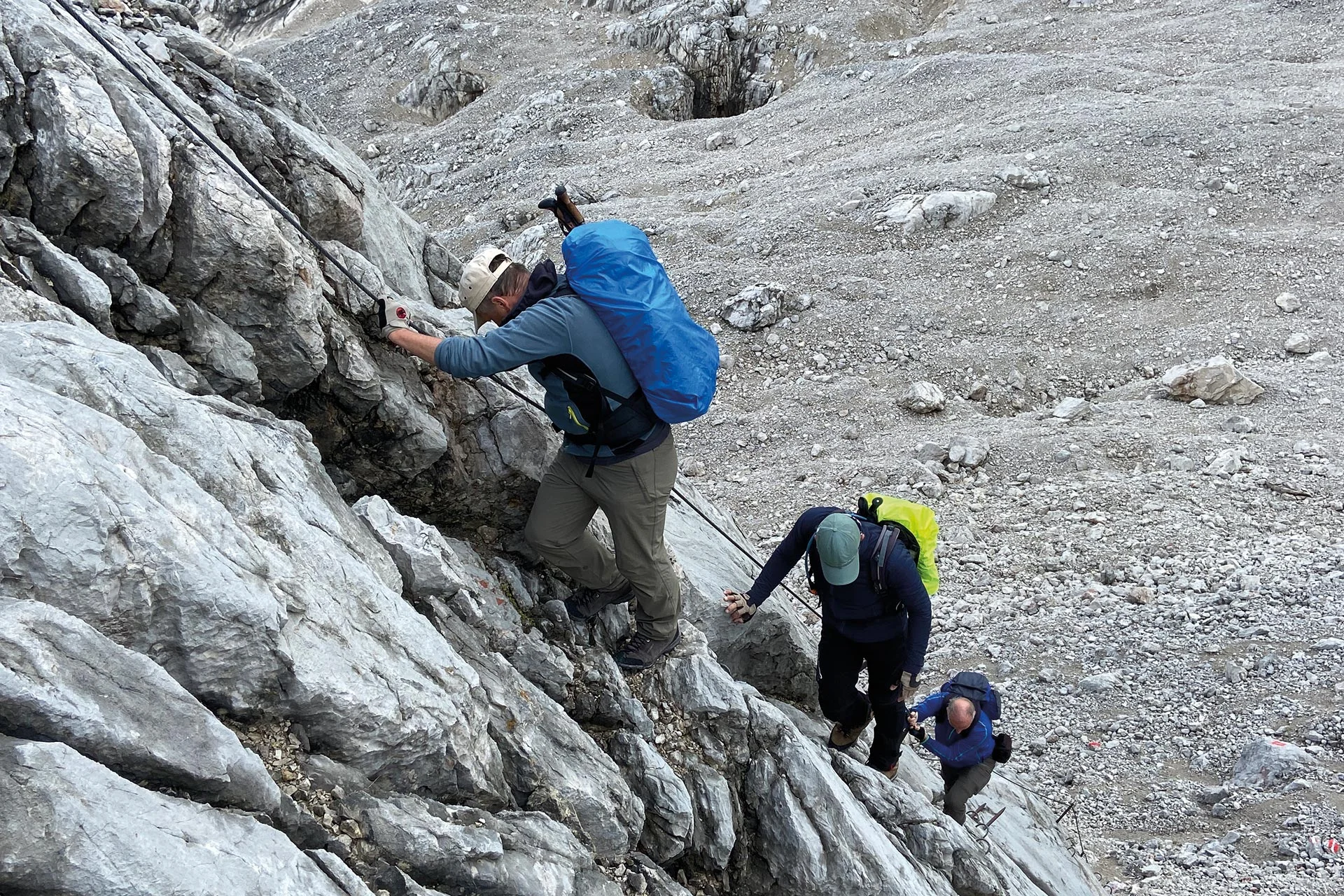 Es sind drei Wanderer mit Rucksäcken zu sehen, die an einem Sicherungsseil nach oben klettern. Alle schauen nach unten oder zur Seite. | © DAV Münster